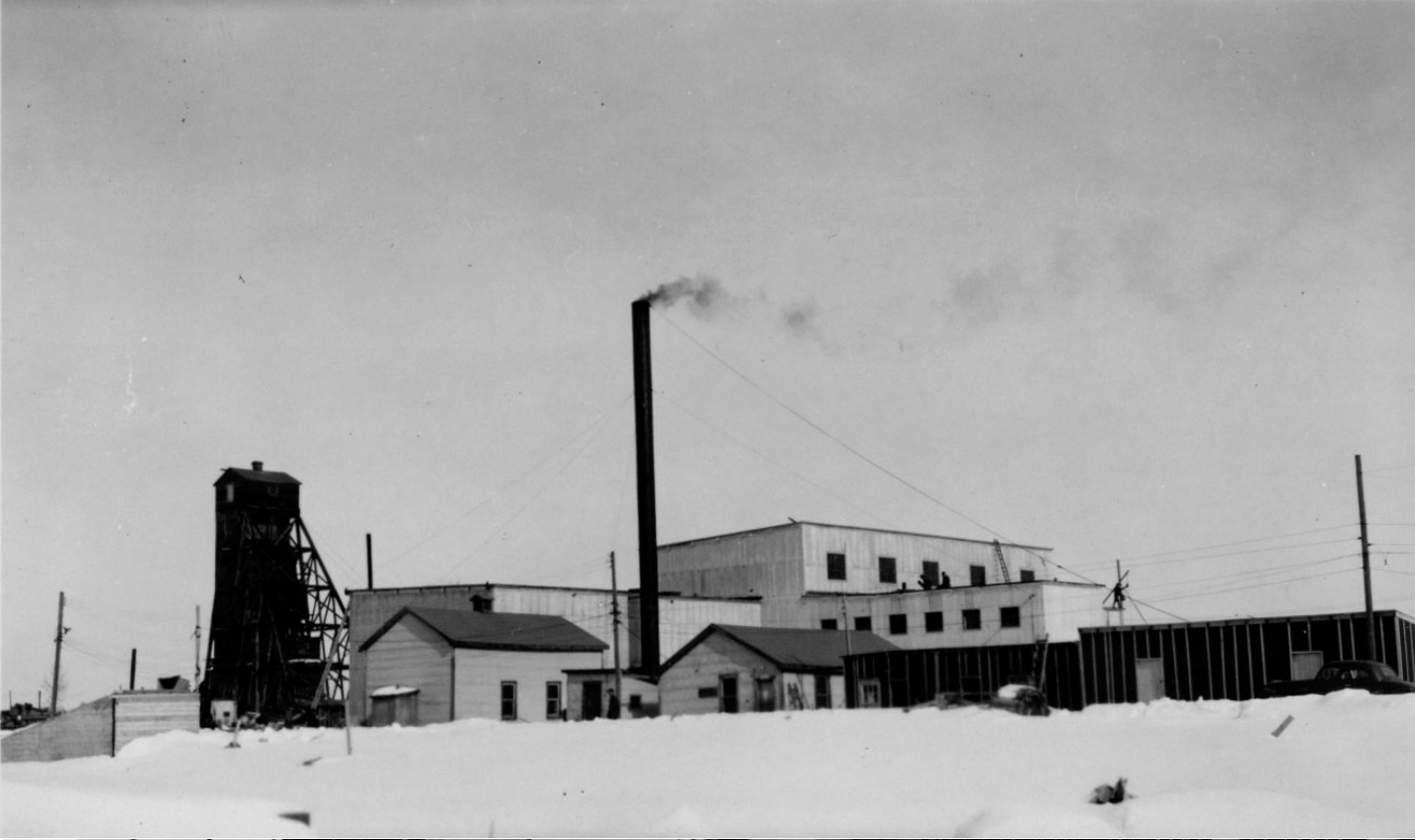 Photographie en noir et blanc de plusieurs bâtiments miniers en planche, dont un chevalement et une cheminée, avec de la neige au sol.
