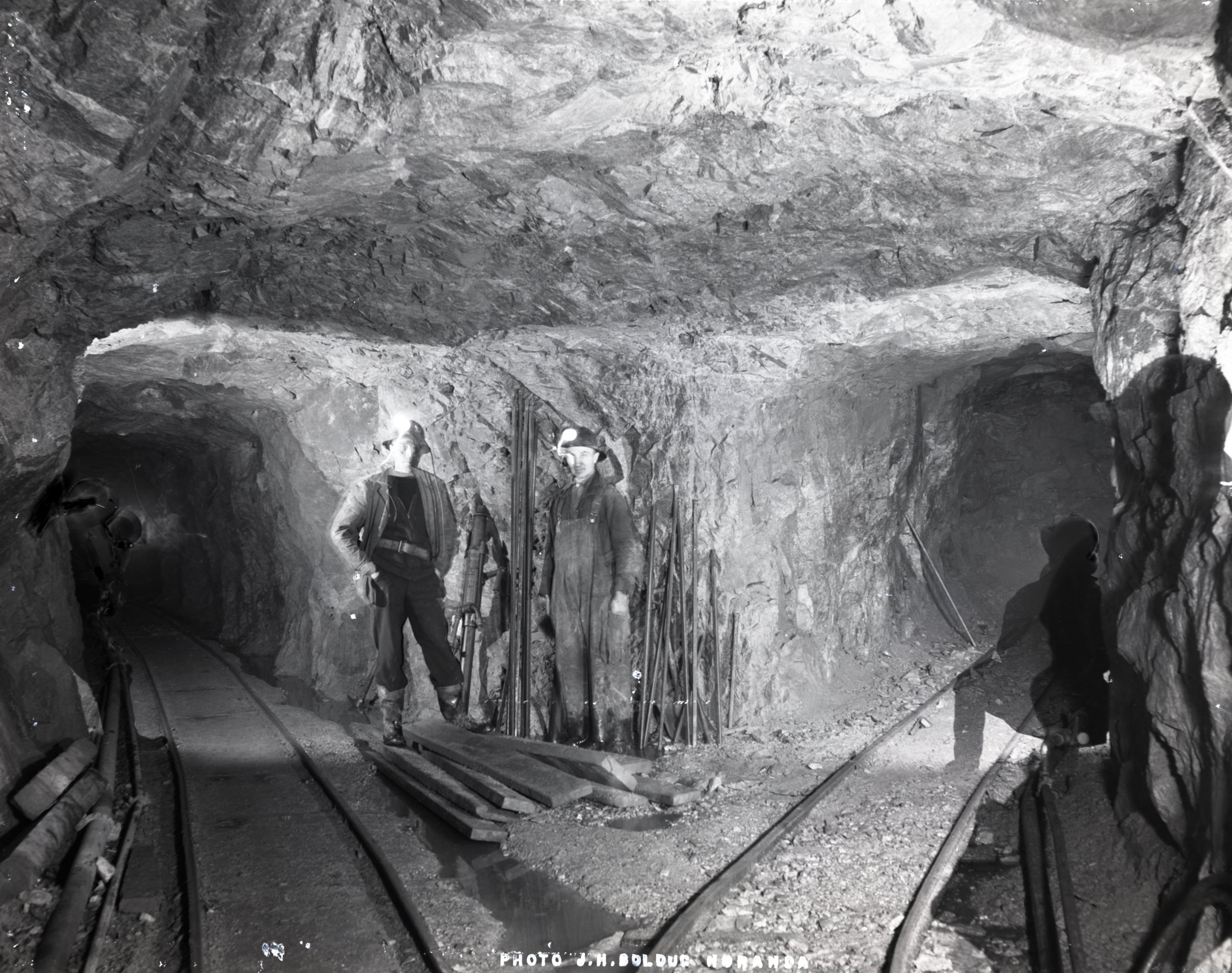 Photographie en noir et blanc de l’intersection entre trois galeries à l’intérieur d’une mine. Il y a deux rails de chemin de fer par terre, ainsi que deux hommes qui posent devant une foreuse et des aciers.