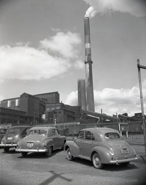 Photographie en noir et blanc  de trois vieilles voitures stationnées devant la clôture de la mine Horne. En arrière-plan, la fonderie ainsi qu'une cheminée qui émet de la fumée.