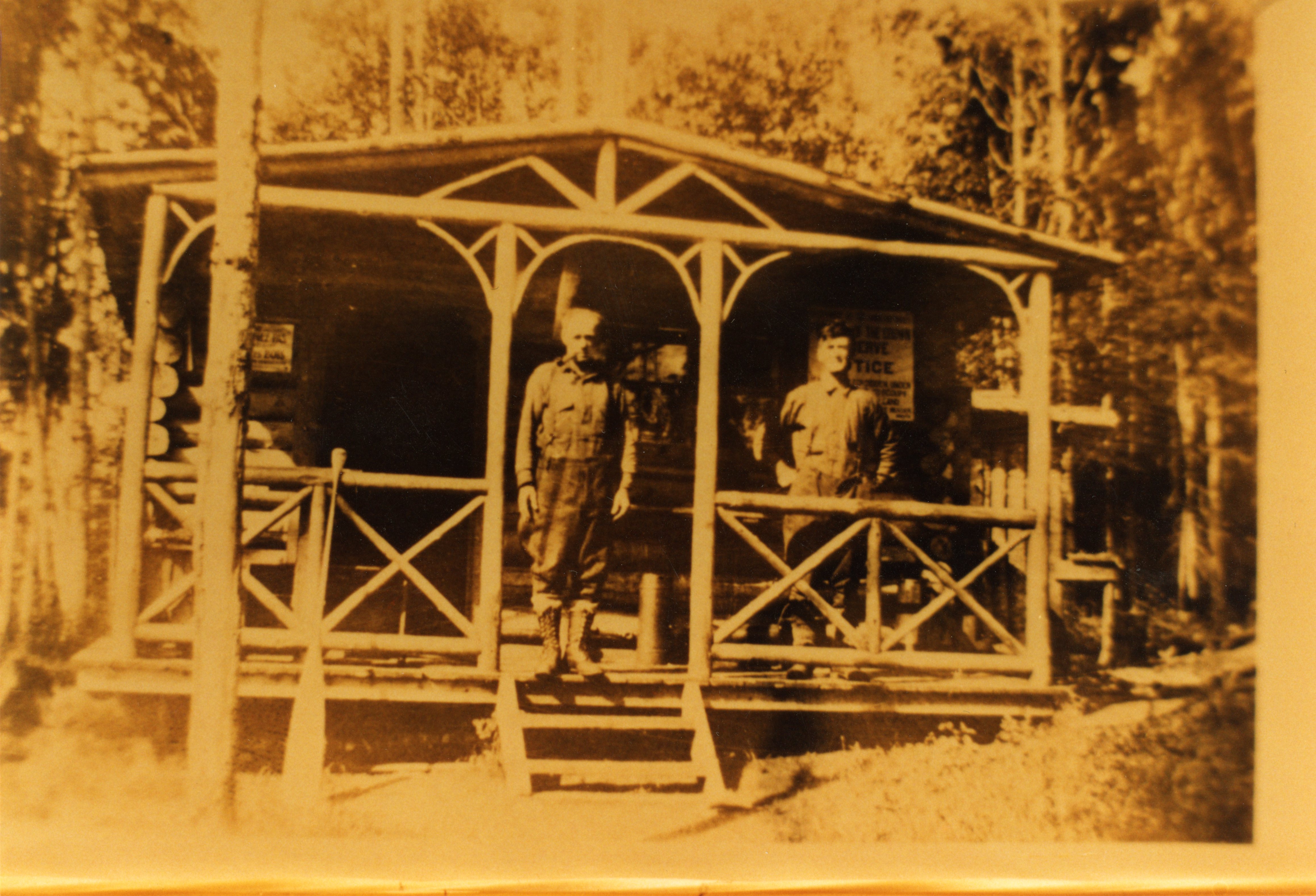 Photographie de couleur sépia de deux hommes debout sur la galerie d'une cabane en bois rond avec la forêt en arrière-plan.