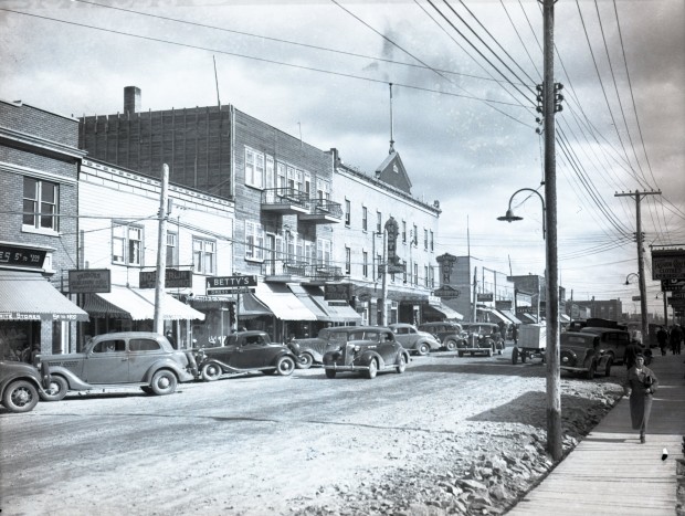 Photographie en noir et blanc d'une rue bordée d'édifice de deux ou trois étages qui ont, pour la plupart, des affiches annonçant des commerces. En plus des voitures qui sont stationnées ou qui  roulent dans la rue, plusieurs personnes circulent sur les trottoirs de bois.