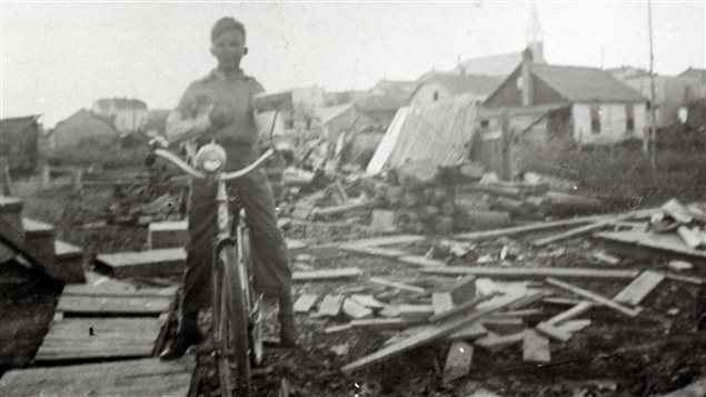 Photographie en noir et blanc d’un petit garçon sur une bicyclette qui pose devant des amoncellements de planches et de buches. En arrière-plan, des maisons et une église. 