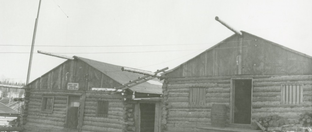 Photographie en noir et blanc de deux cabanes en bois rond avec des barreaux d’installés sur les fenêtres. Une affiche est posée au-dessus de la porte du bâtiment de gauche.