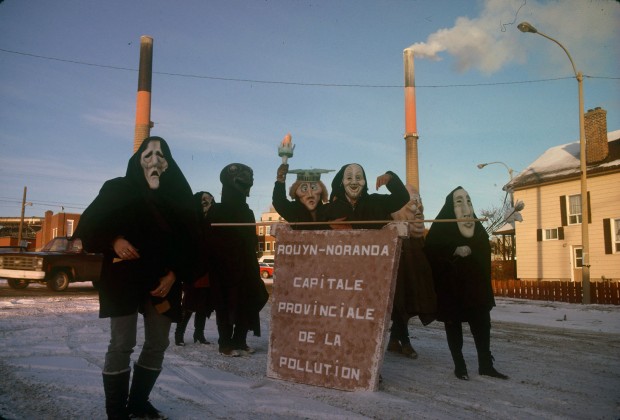 Photo couleur d'un groupe de sept personnes, devant la fonderie Horne, qui sont déguisé et qui transporte une affiche qui porte l'inscription : Rouyn-Noranda capitale provinciale de la pollution.