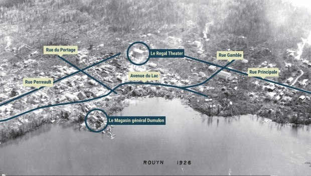 Photo noir et blanc, prise à bord d'un avion, du village naissant de Rouyn situé sur la rive sud du lac Osisko. Le tracé et le nom des principales artères sont identifiés, Rue du Portage, Perreault, Gamble, Principale et Avenue du Lac. Deux commerces importants, Le Régal Theater et Le Magasin général Dumulon, sont identifiés.