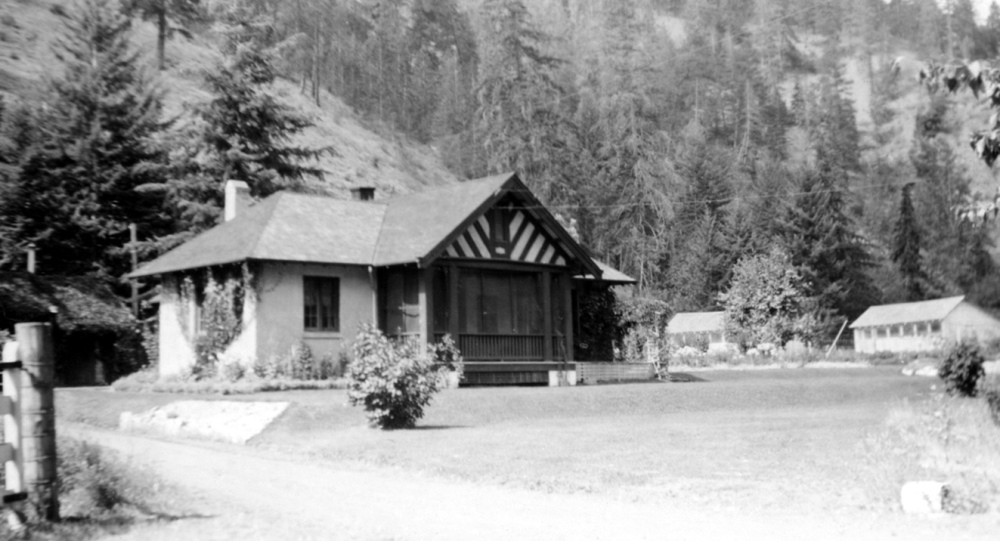 Photo en noir et blanc montrant une maison à pignons, deux bâtiments plus petits à droite et une colline à l’arrière-plan.