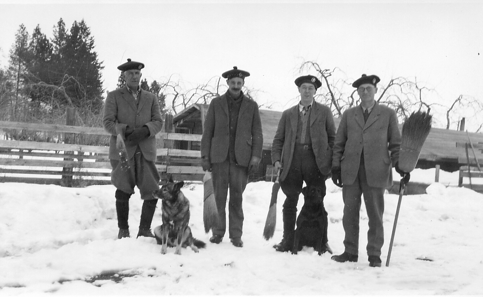 Photo en noir et blanc montrant quatre hommes debout dans la neige tenant des balais. Les hommes portent tous des bérets et des vestes de costume. Un chien est assis près d’eux.