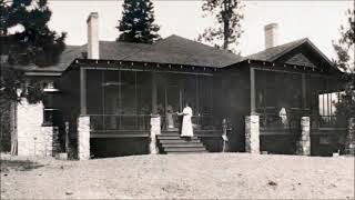 Photo en noir et blanc montrant une maison entourée d’une véranda. Une femme en robe blanche se tient debout sur les marches.