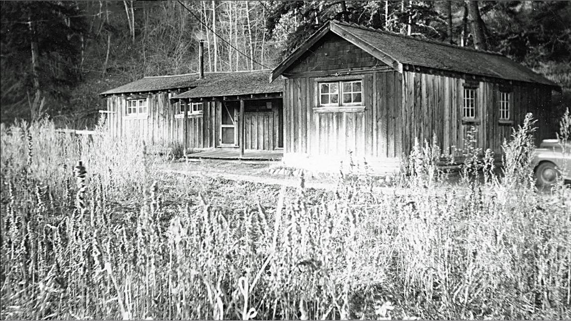 Photo en noir et blanc d’un bâtiment en bois. De hautes herbes occupent le premier plan.