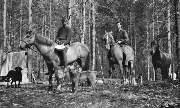 Photo en noir et blanc montrant deux hommes à cheval dans une forêt. Un troisième cheval et deux chiens les accompagnent.
