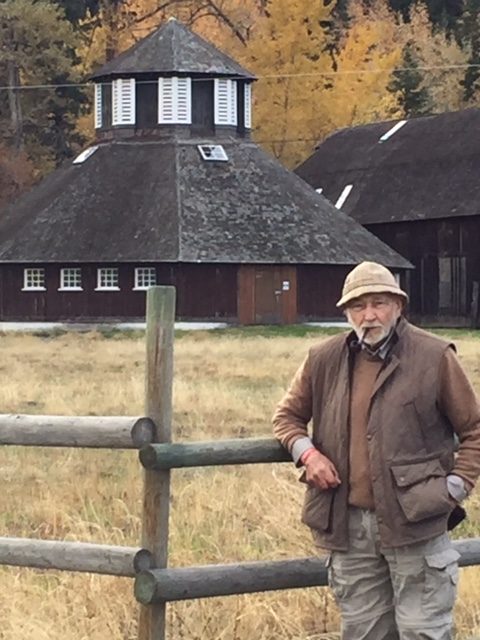 Photo en couleur montrant un homme avec un chapeau et une pipe, appuyé sur une clôture près d’une étable ronde.