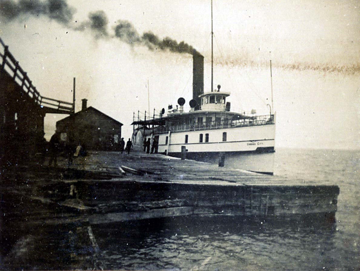 Photographie en noir et blanc d'un bateau à vapeur amarré au quai. Petit bâtiment sur la jetée à gauche du bateau, et un petit groupe de personnes sur la jetée à côté du bateau.
