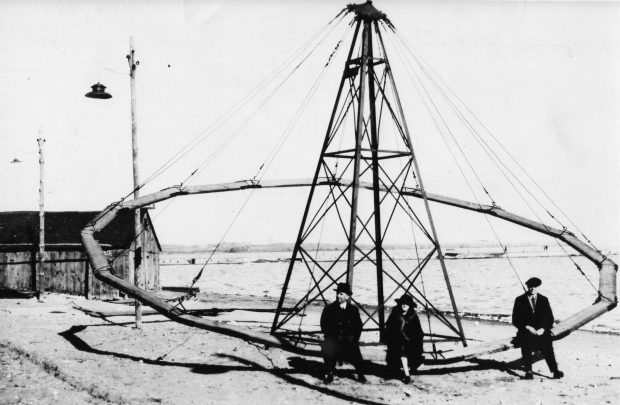 Photographie en noir et blanc de trois personnes assises sur une balançoire circulaire le lac et la jetée et sont à l'arrière-plan.