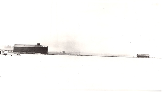 Photographie en noir et blanc d'un grand hangar de stockage situé à gauche de la jetée menant à un plus petit hangar.