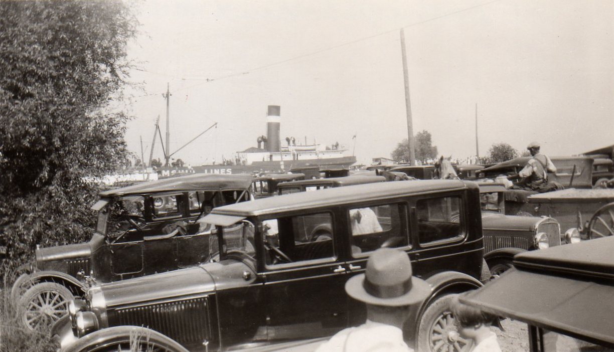 Photographie en noir et blanc de vieilles voitures garées avec des gens debout autour d'eux. Un bateau à vapeur est visible en arrière-plan.