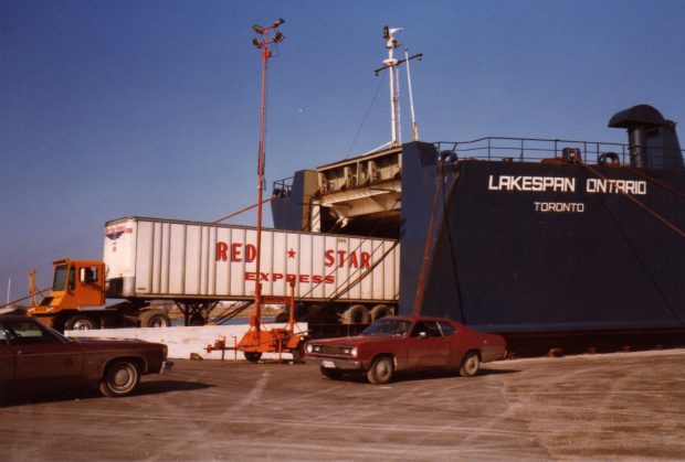 Photographie couleur d'un camion de transport rouge et blanc reculant dans un gros navire, Lakespan Ontario, amarré au quai.