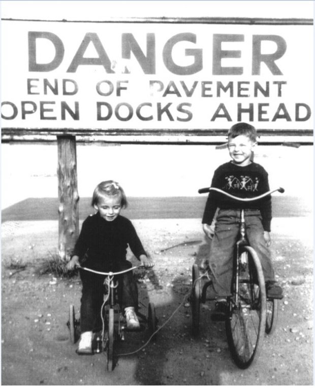 Photographie en noir et blanc de deux enfants sur des tricycles près d'un grand panneau indiquant Danger End of Pavement Open Docks Ahead.