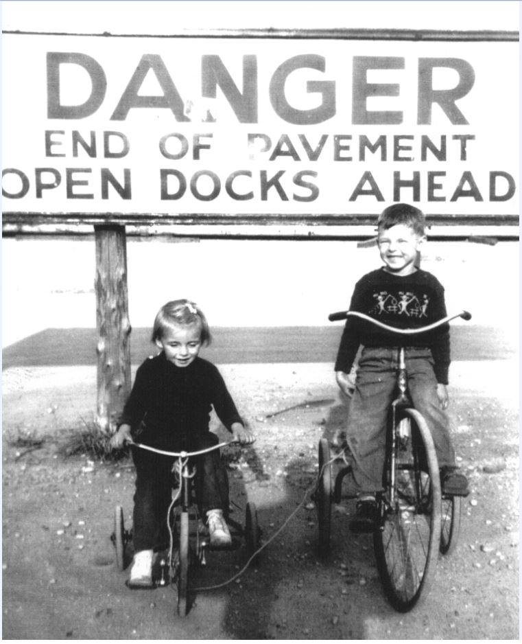 Photographie en noir et blanc de deux enfants sur des tricycles près d'un grand panneau indiquant "Danger End of Pavement Open Docks Ahead".