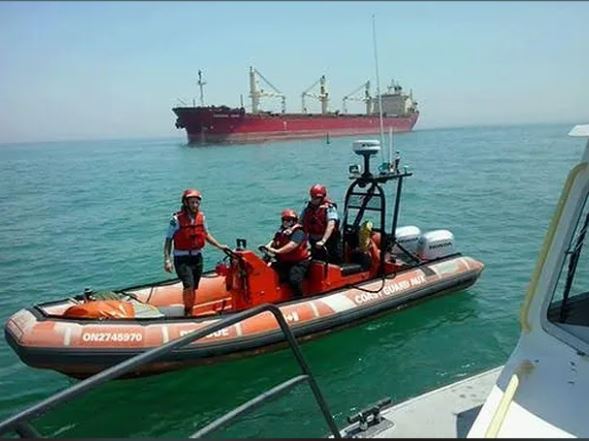 Photographie couleur de trois personnes portant des casques et des gilets de sauvetage dans un petit bateau de sauvetage orange de la Garde côtière sur l'eau. Plus grand navire rouge en arrière-plan.
