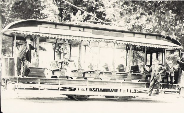 Photographie en noir et blanc d'un tramway avec côtés ouverts et bancs au milieu. Trois personnes debout et appuyées sur le tramway et une personne assise sur le siège.