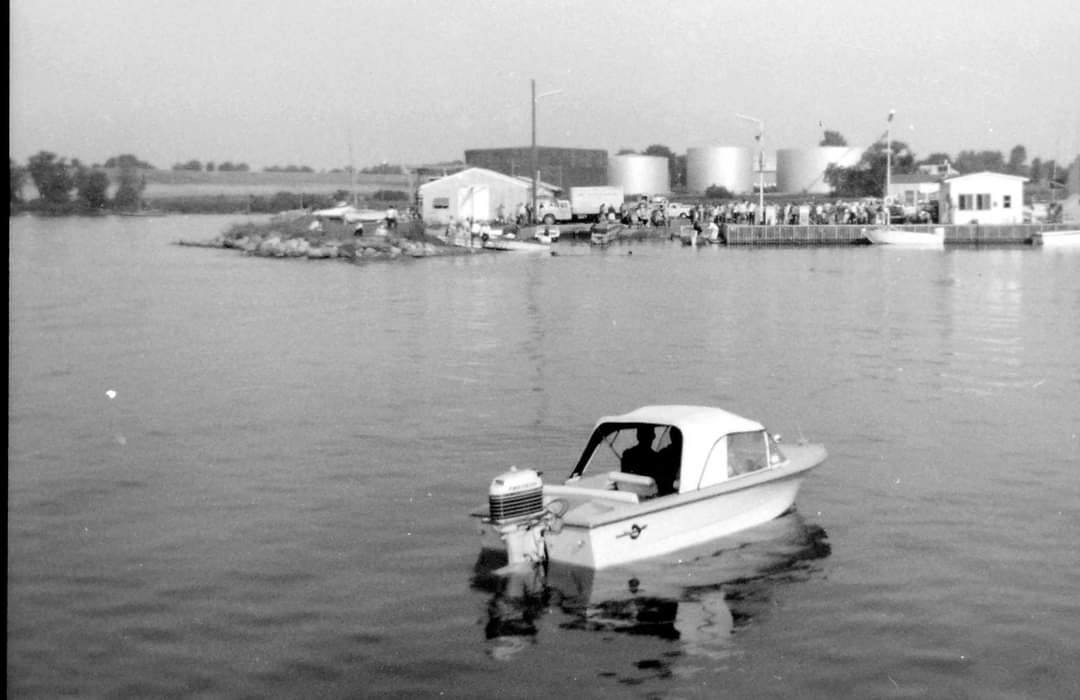 Photographie en noir et blanc d'un petit bateau à moteur sur l'eau. En arrière-plan, des bâtiments, des bateaux, des quais et des réservoirs de pétrole.