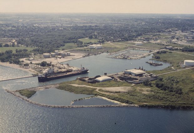  Photographie aérienne en couleur des terres du port d'Oshawa. Il y a un grand navire amarré dans le chenal principal, quelques bâtiments portuaires au centre de l'image et une vue sur la marina en arrière-plan.