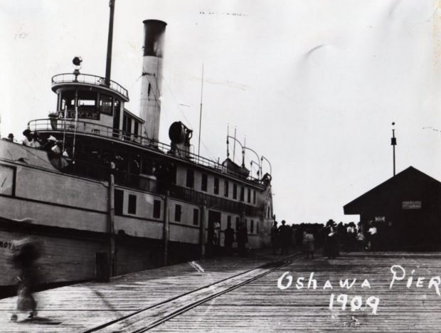Photographie en noir et blanc d'un bateau à vapeur amarré au quai. Petit bâtiment sur la jetée à droite du bateau et grand groupe de personnes debout sur la jetée à côté du bateau.