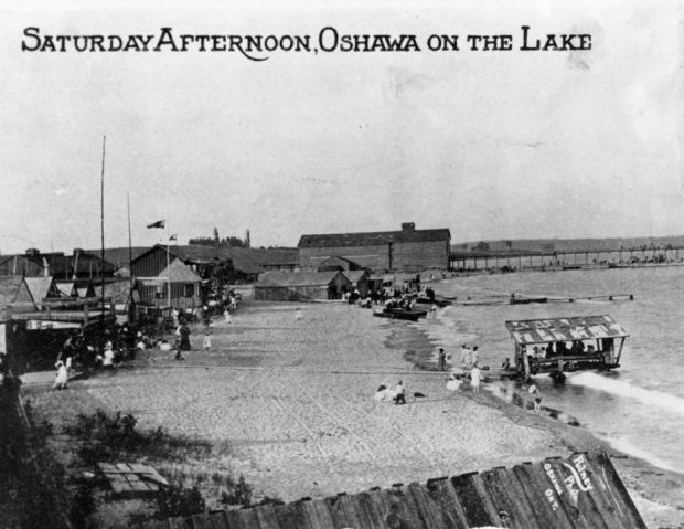 Black and white post card of several people in suits and long skirts on a beach. There are some small buildings on the left, and a wooden building leading into a long, wooden pier in the background.