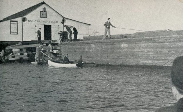 Photographie en noir et blanc d'un grand bâtiment de yacht club. Un groupe de personnes est autour du bâtiment et un plus petit bateau est dans l'eau.