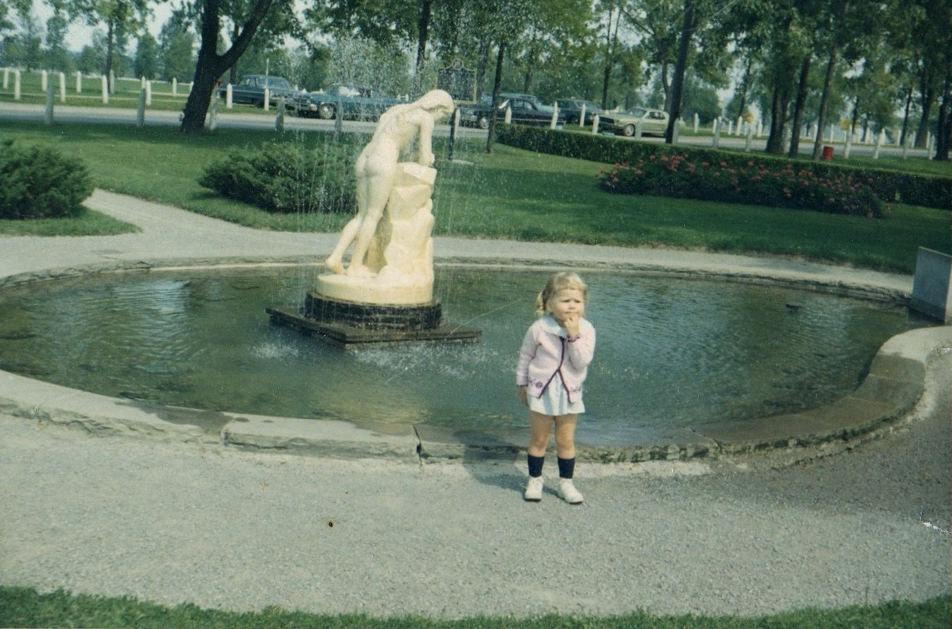 Photographie couleur d'un enfant debout devant une statue de marbre entourée d'un bassin d'eau. Des arbres bordent la rue derrière la statue et des voitures sont garées dans la rue.