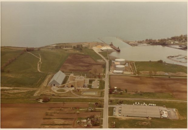 Photographie aérienne en couleur des terres entourant le port d'Oshawa. Il y a deux grandes usines et un bateau décharge le sucre brut au port.