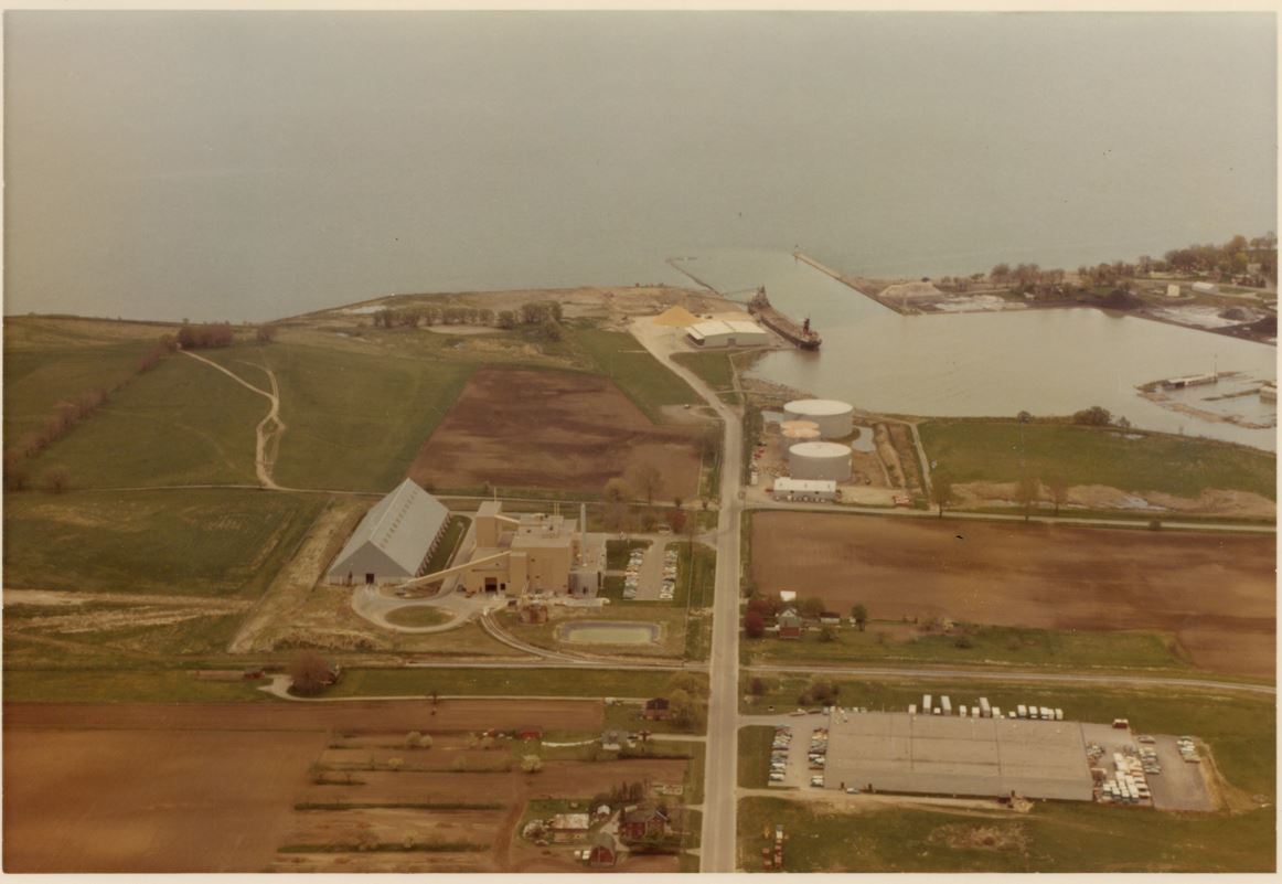 Photographie aérienne en couleur des terres entourant le port d'Oshawa. Il y a deux grandes usines et un bateau décharge le sucre brut au port.