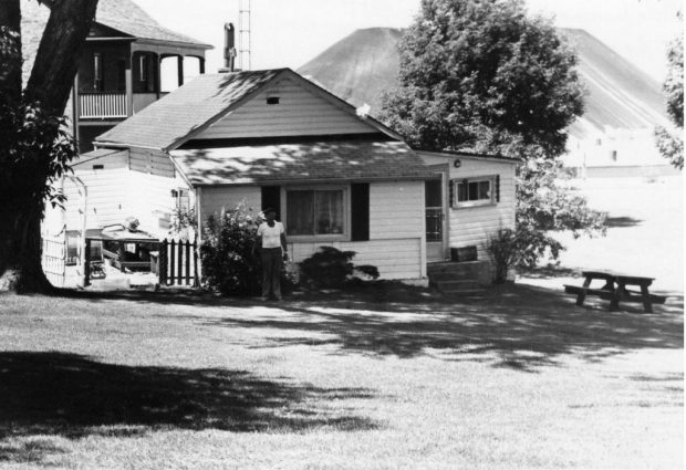 Photographie en noir et blanc d'un petit chalet, d'une maison plus grande derrière et de gros tas de charbon à l'arrière-plan.