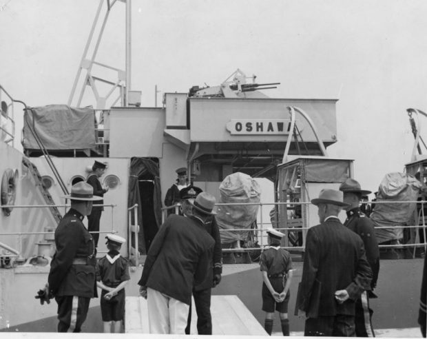 Photographie en noir et blanc d'un bateau, H.M.C.S. Oshawa, avec des gens en uniformes et costumes sur le pont du bateau et debout sur la jetée à côté du bateau.