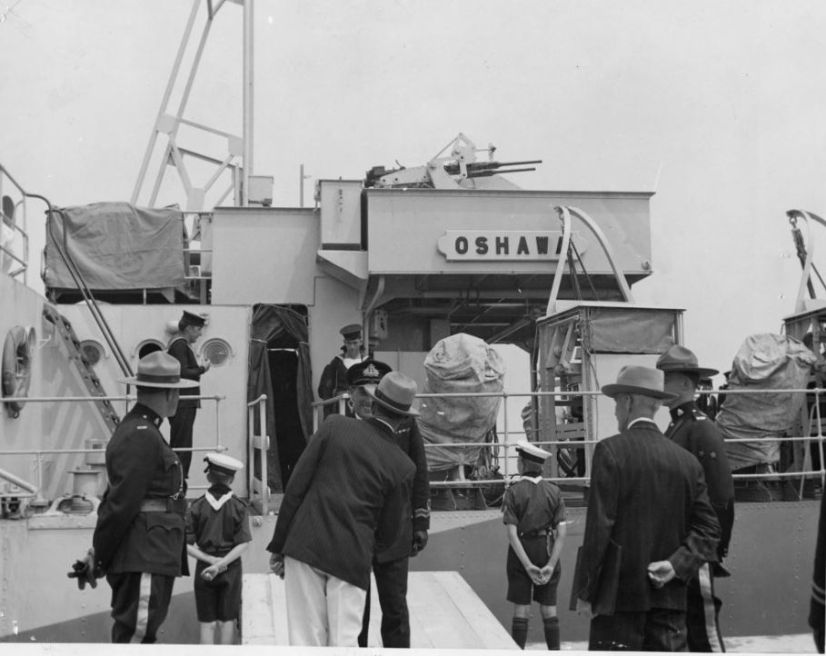 Photographie en noir et blanc d'un bateau, H.M.C.S. Oshawa, avec des gens en uniformes et costumes sur le pont du bateau et debout sur la jetée à côté du bateau.