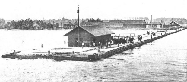 Copie en noir et blanc des gens et d'un petit bâtiment debout sur une jetée en bois. D'autres bâtiments sont en arrière-plan.