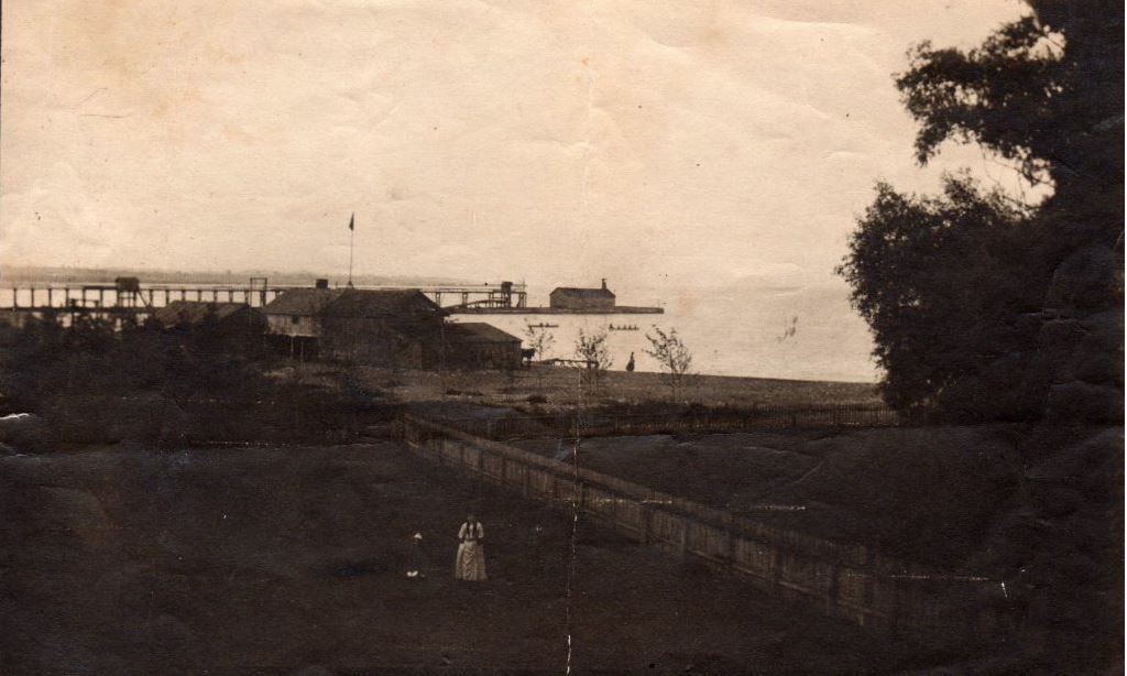Photographie en noir et blanc d'une personne dans un champ avec de grands bâtiments au loin. La jetée et le lac sont visibles en arrière-plan, derrière les bâtiments.