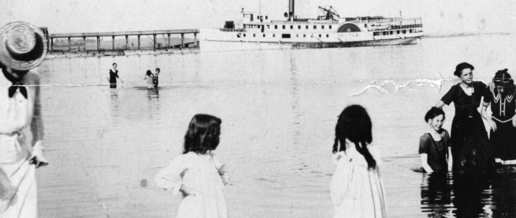 Photographie en noir et blanc d'un lac avec des individus debout dans l'eau et à l'arrière-plan un grand bateau à vapeur et une jetée