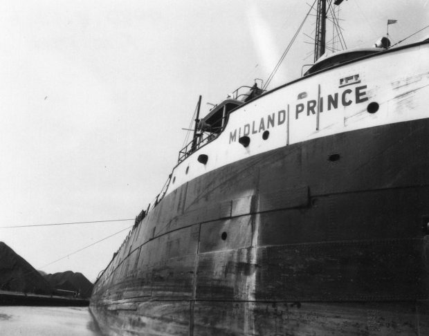 Photographie en noir et blanc d'un grand navire. Midland Prince est écrit sur le côté du navire. Les tas de charbon sont en arrière-plan.