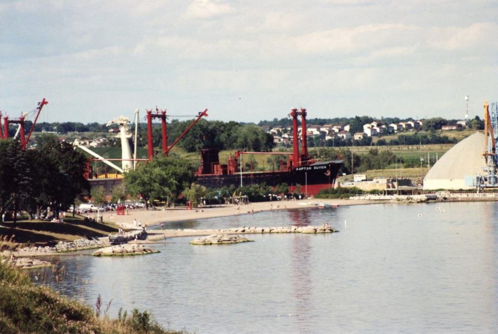 Photographie couleur d'une plage avec quelques arbres et un grand navire amarré au port d'Oshawa derrière.