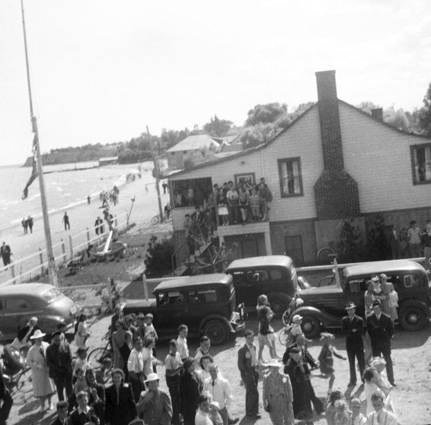 Photographie en noir et blanc d'un groupe de personnes au bord du lac. Des voitures et un bâtiment avec une grande ancre devant.