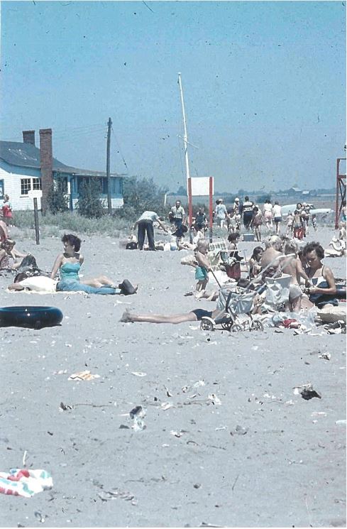 Photographie couleur de plusieurs personnes assises sur la plage avec une maison en arrière-plan.