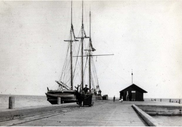 Photographie en noir et blanc d'une goélette amarrée au quai avec un petit bâtiment à droite du quai. Il y a une personne avec un cheval et une charrette à côté de la goélette sur la jetée.