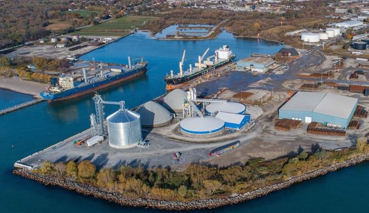 Photographie aérienne en couleur du port d’Oshawa. Deux grands bateaux sont amarrés au chenal au milieu de l’image, et plusieurs bâtiments industriels se trouvent à l’arrière-plan, ainsi qu’une plage et des arbres de l’autre côté du port.  