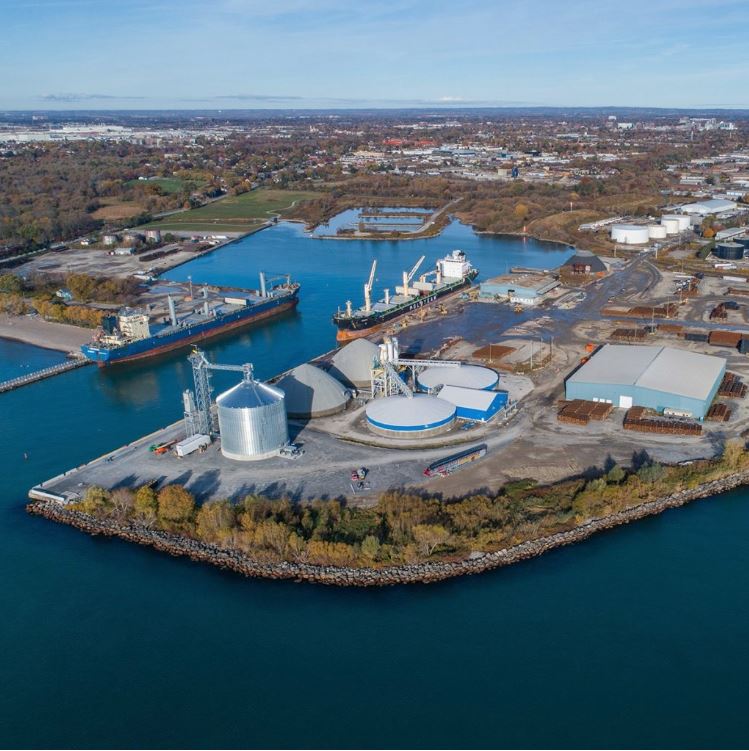 Photographie aérienne en couleur du port d’Oshawa. Deux grands bateaux sont amarrés au chenal au milieu de l’image, et plusieurs bâtiments industriels se trouvent à l’arrière-plan, ainsi qu’une plage et des arbres de l’autre côté du port.  