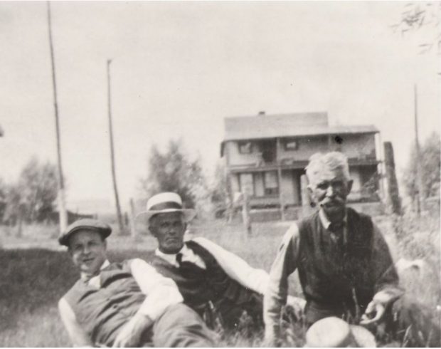Photographie en noir et blanc des trois personnes assises dans l'herbe. Une grande maison à deux étages est à l'arrière-plan.