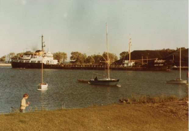  Photographie couleur d'un grand navire amarré au port déchargeant du charbon. Une personne est vue près du bas de l'image et de petits bateaux sont ancrés dans l'eau.