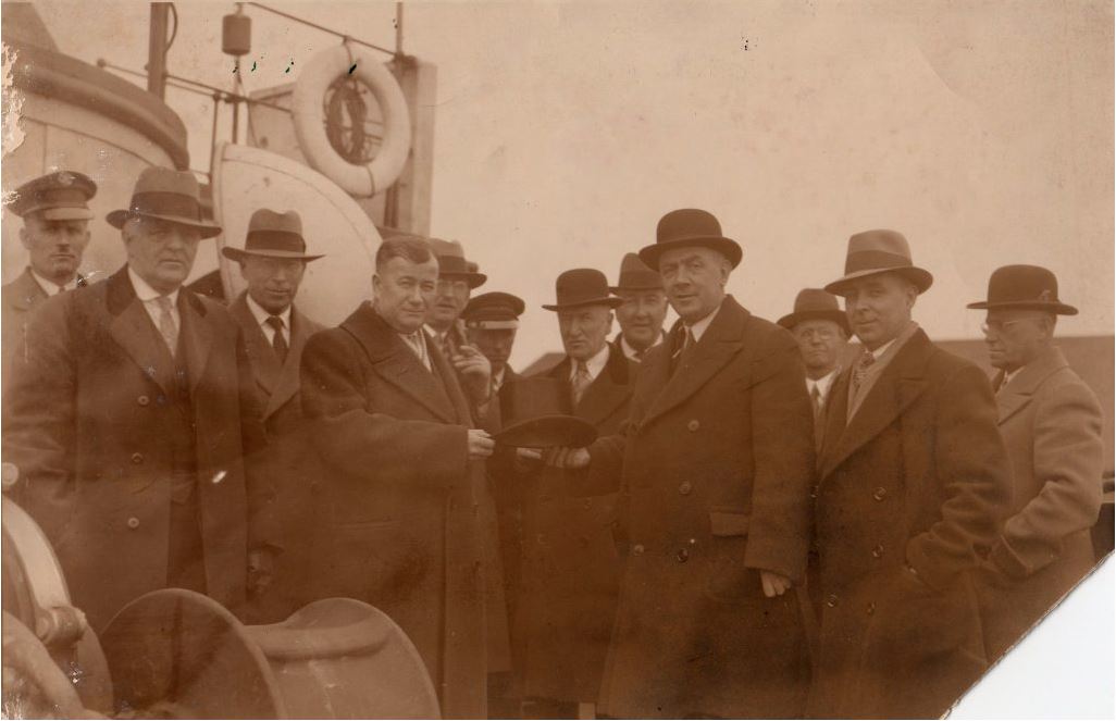 Sepia photograph of a group of people in coats and hats. Person at front right is giving a top hat to person at front left.