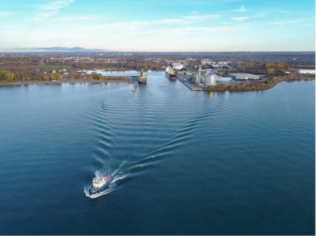 Photographie aérienne en couleur d'un port avec deux navires amarrés aux quais est et ouest. Un remorqueur quitte le port.