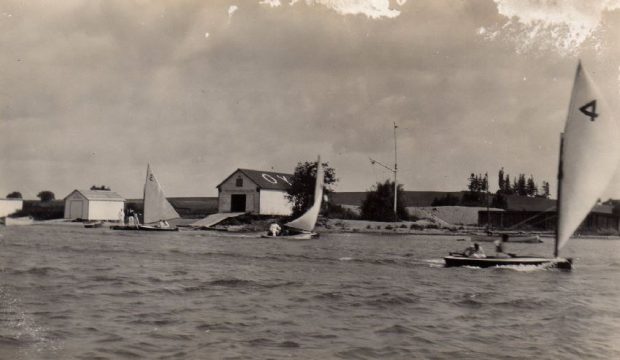 Photographie en noir et blanc d'un grand bâtiment de yacht club avec deux bâtiments plus petits sur la droite. Les trois bâtiments sont situés le long du lac.